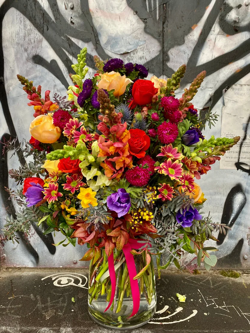 Vibrant vase arrangement of orange roses, purple anemones and snapdragons with a magenta ribbon against a graffiti wall