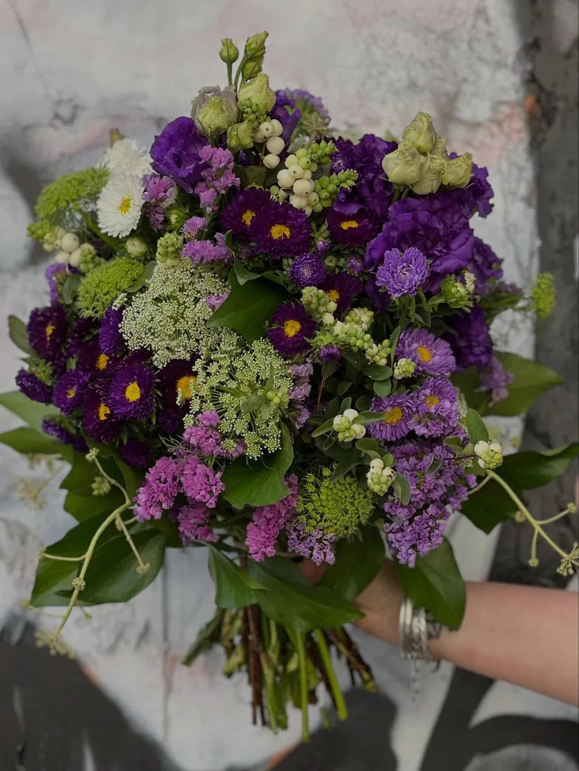 Wild Purple Posy — purple asters, statice, lisianthus, snowberry and ivy held against a pale graffiti wall