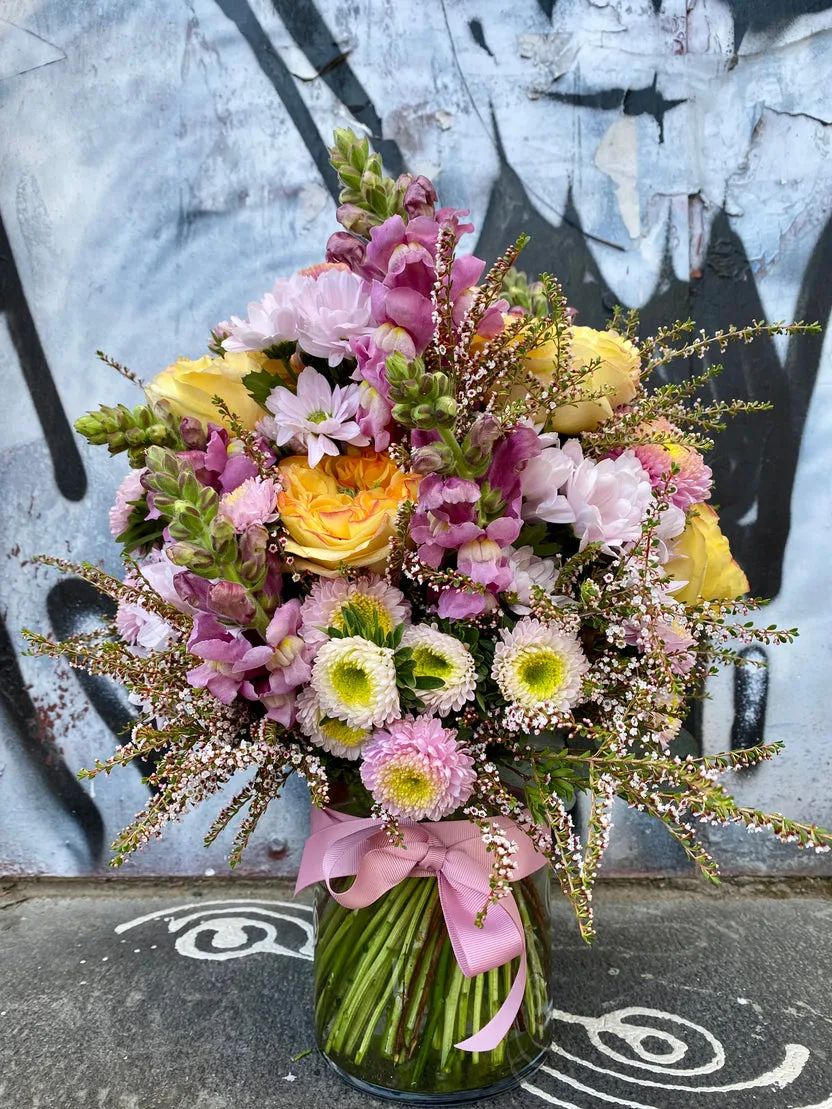 Pastel vase arrangement with pink snapdragons and butter-yellow roses, tied with ribbon, photographed against Smith Street's graffiti walls