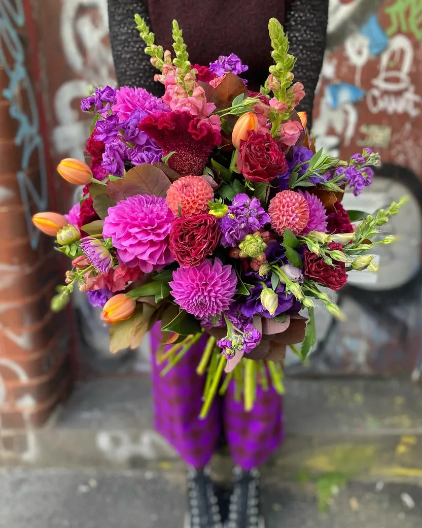 Statement bouquet — magenta dahlias, crimson roses and purple stock held against a graffiti wall